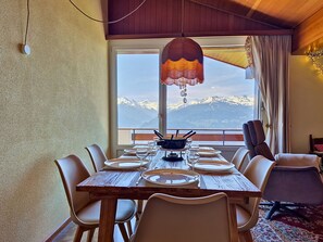 Ein Essbereich mit einem Holztisch, gedeckt für vier Personen, mit Blick auf verschneite Berge durch ein großes Fenster und einer hängenden Leuchte.