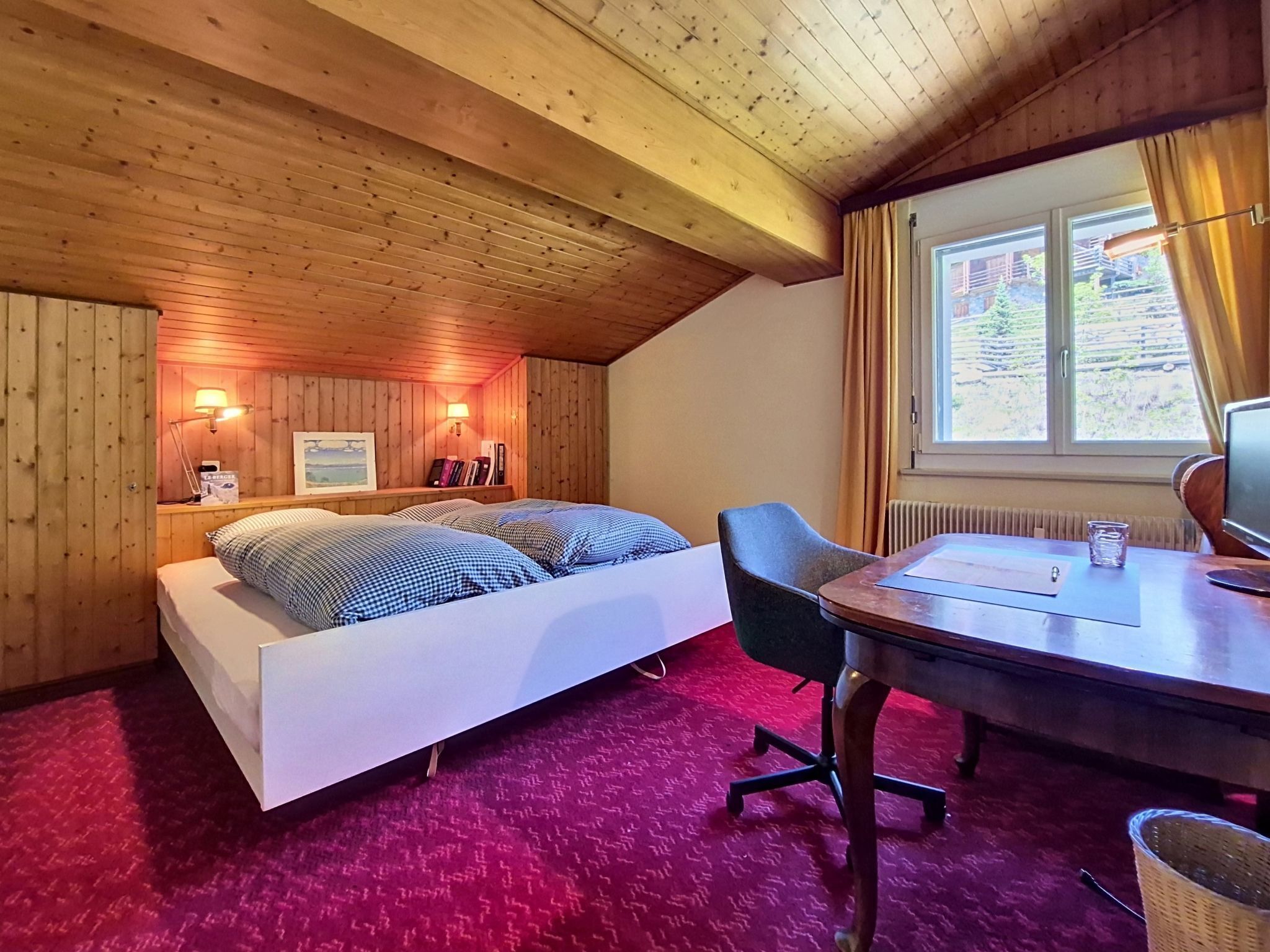 Ein Zimmer mit Holzboden, einem Bett mit kariertem Bettzeug, einem Schreibtisch mit Stuhl und einem Fenster mit Vorhängen.