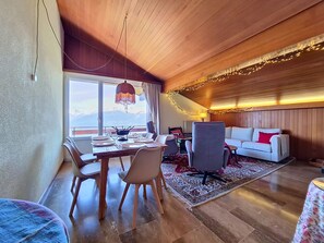 Ein Essbereich mit Tisch für vier Personen, einer Couch, einem gemusterten Teppich und Blick auf Berge durch große Fenster.