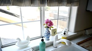 Une cuisine avec une fenêtre, un évier blanc, un flacon de liquide vaisselle, un vase avec des fleurs et un distributeur de savon.