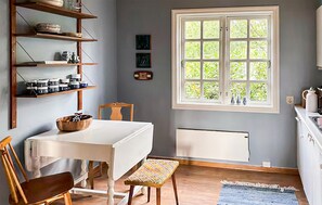 Een eetruimte met een tafel, stoelen, een bank, een raam met uitzicht op groen en planken met keukengerei.