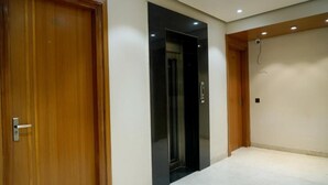 Habitación de hotel con puerta de madera, un ascensor negro y una alfombra blanca.