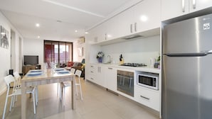 A modern kitchen with white cabinetry, a stainless steel refrigerator, a built-in oven, and a dining area with a table and chairs. - Modern 2BR 3 min to Central Station FREE Parking (Chippendale)