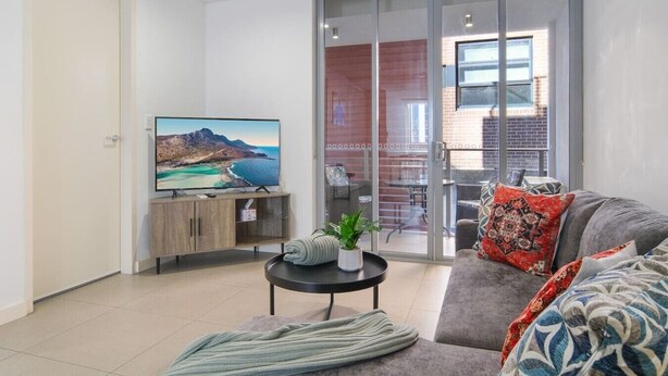 A modern living room with a flat-screen TV, a sofa, a coffee table, and a sliding glass door leading to an outdoor area. - Modern 2BR 3 min to Central Station FREE Parking (Chippendale)