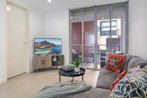 A modern living room with a flat-screen TV, a sofa, a coffee table, and a sliding glass door leading to an outdoor area. - Modern 2BR 3 min to Central Station FREE Parking (Chippendale)