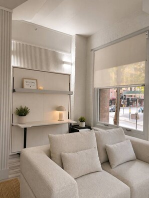 A modern living room with a sofa, a white console table with decorative items, and a large window offering a view of the street. - Classic City Unit with Parking and walk to Harbour (Pyrmont)