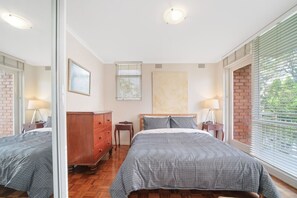 A bedroom with two beds, a dresser, a bedside table, a lamp, a window with blinds, and a framed picture on the wall. - Cozy 1 Bed Harbour View Apartment and Free Carpark (North Sydney)