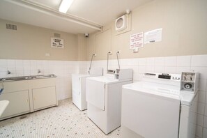 Ein Waschraum mit drei weißen Waschmaschinen, einem Waschbecken sowie zwei Schildern mit Angaben zu den Waschzeiten und der Nutzung eines Ventilators.
