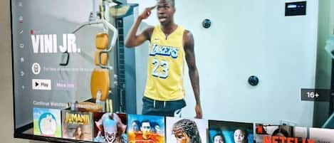 A television mounted on a wall displaying a sports figure in a yellow jersey, with a remote control, a lamp, and a small plant on a table below.