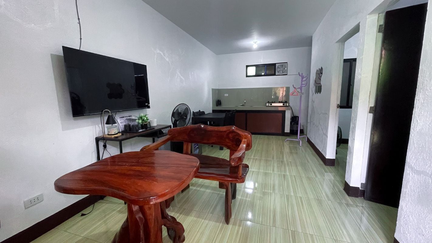 A dining area with a wooden table and chairs, a flat-screen TV mounted on the wall, and a kitchen in the background.