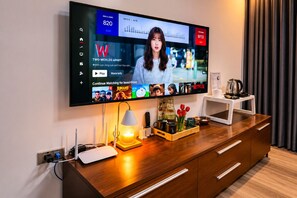 Un televisor de pantalla plana montado en la pared mostrando un noticiero, con un escritorio de madera debajo que tiene una lámpara, una cafetera y una bandeja con objetos.
