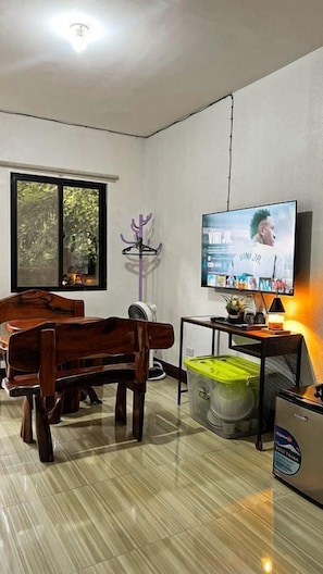A room with a wooden dining table, a television mounted on the wall, a small table with a lamp, and a trash bin. - Upgrade Beach Resort Infanta Quezon (Infanta)