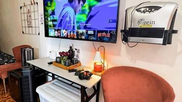 A room with a television mounted on the wall, a small table with a lamp, a chair, and a portable heater.