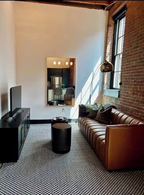 A modern living room with a sofa, a coffee table, a TV, and a kitchen area visible through a glass door. - Brick & Bed - Arch, Casino, Convention Center, T-410 (St. Louis)