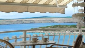 Ein Balkon mit Korbstühlen und einem kleinen Tisch, der Blick auf die Küste mit einer Umzäunung und einer fernen Uferlinie gewährt.