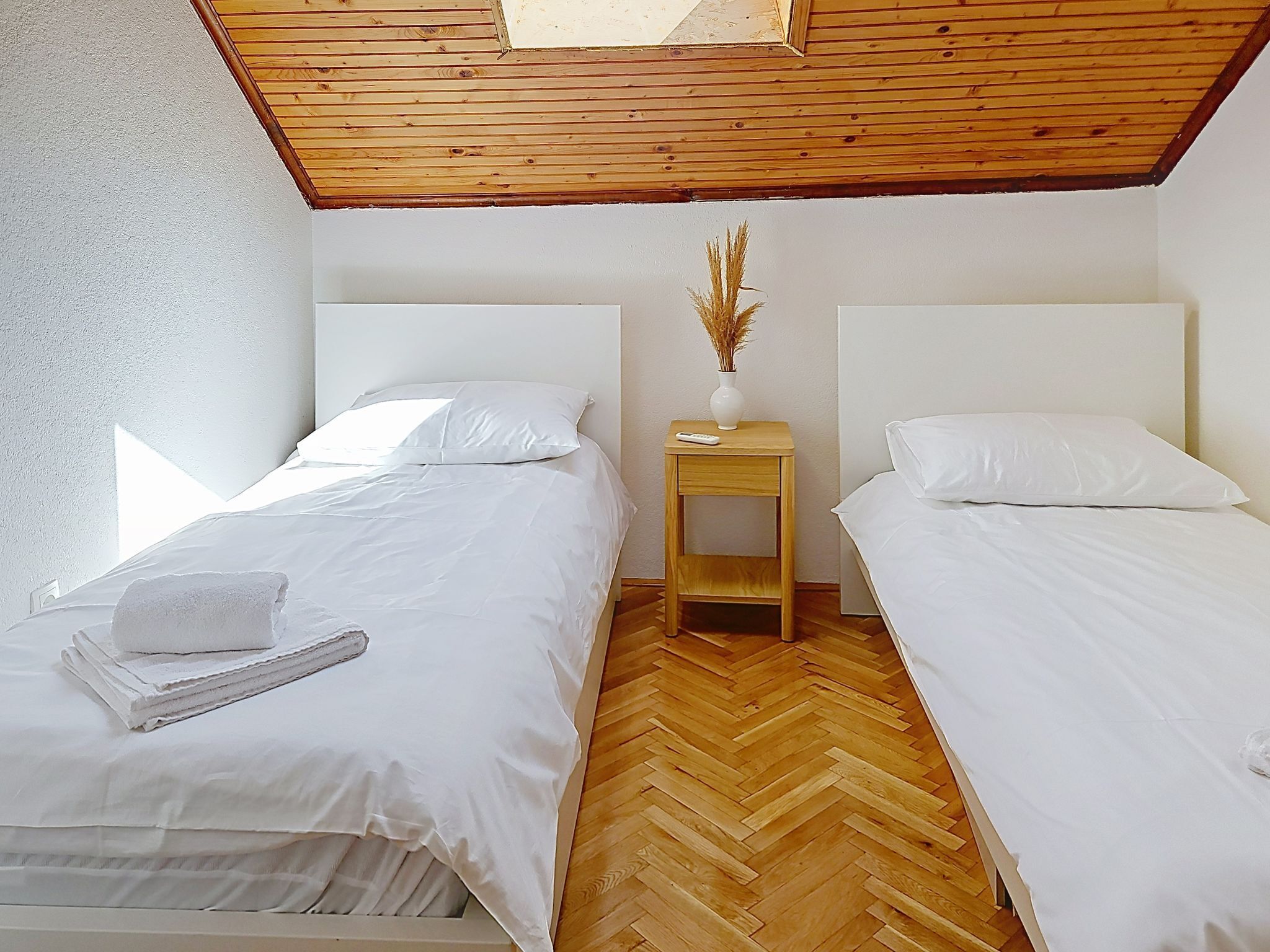 Un dormitorio con dos camas, suelo de madera, un tragaluz y una mesita de noche con un florero.