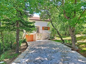 Ein Steinweg führt zu einem Haus mit einer Ziegelwand und einer Holztür, umgeben von Bäumen.