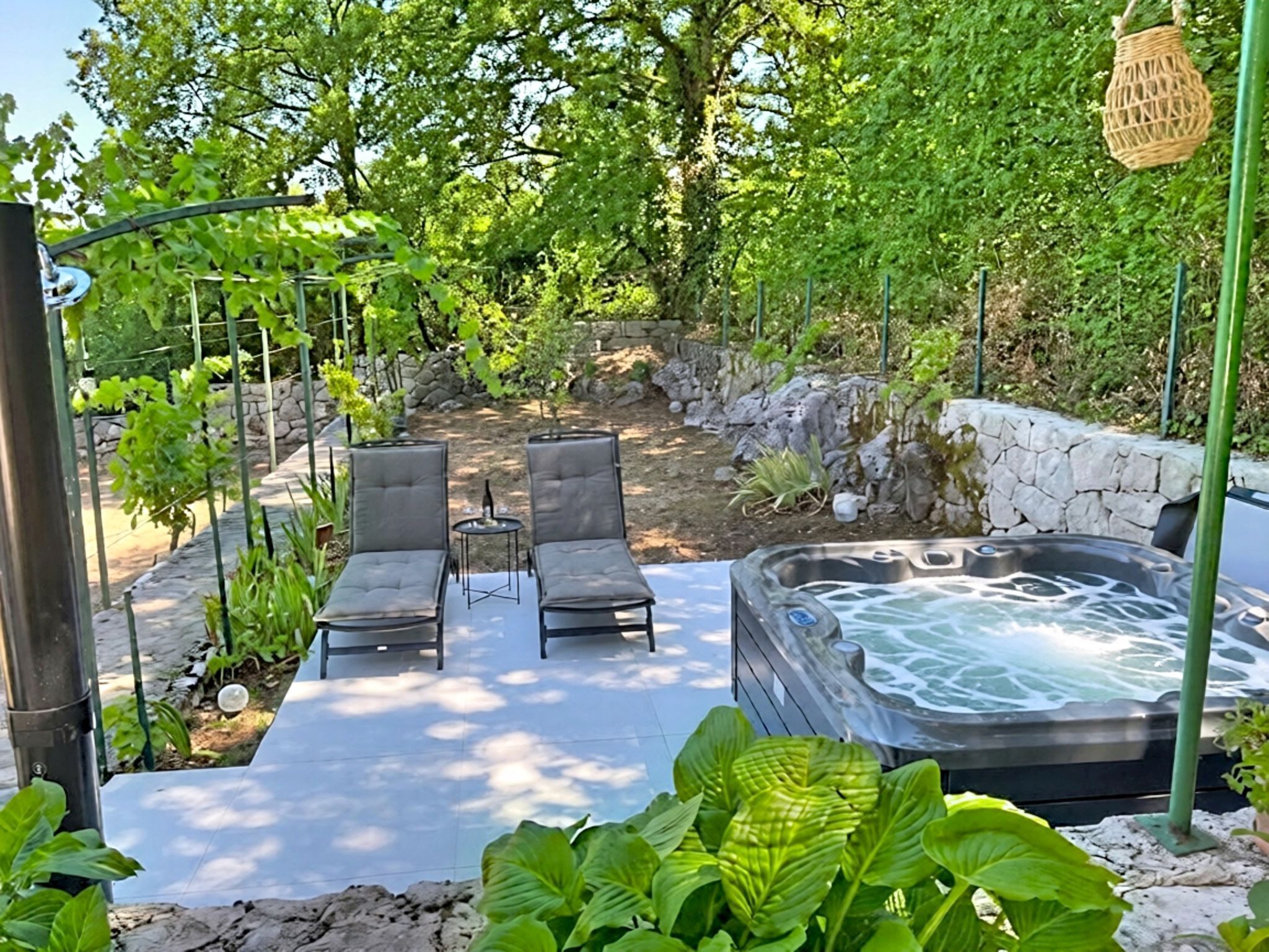 Un bain à remous entouré de chaises longues et de verdure, avec un mur de pierre et une pergola en arrière-plan.