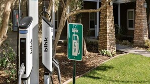 En blink EV-ladestation med et grønt skilt, en murstensbygning og et træ i baggrunden.