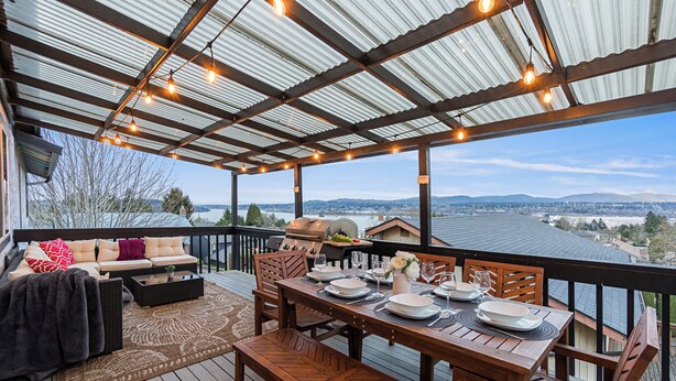 A covered deck with a dining table set for four, outdoor furniture, and a view of a cityscape. - Rainer Estate (Seattle)