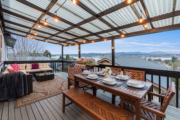 A covered deck with a dining table set for four, outdoor furniture, and a view of a cityscape. - Mins to Seattle~Lake Views~Movie Room/Game Room (Seattle)
