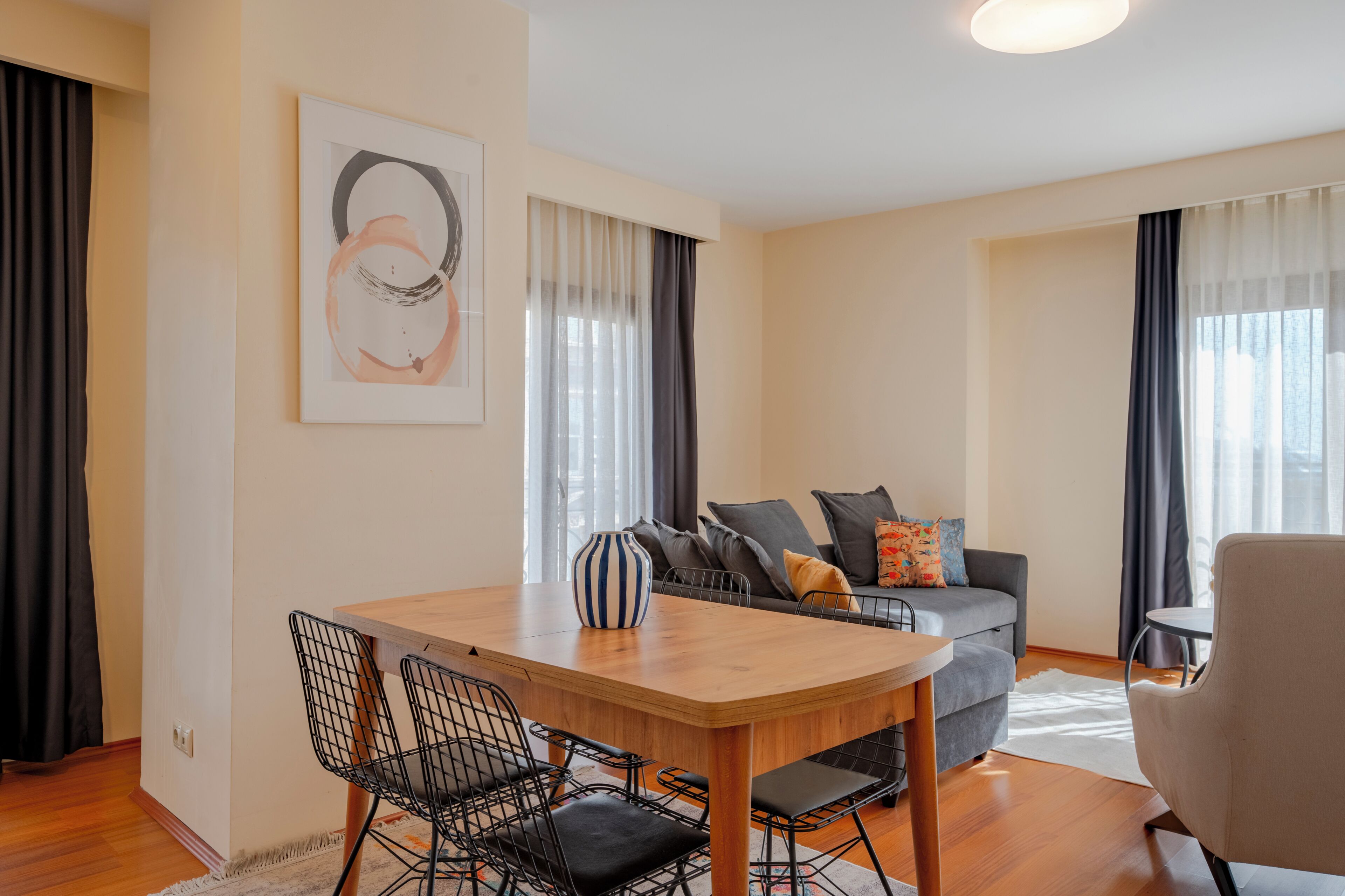 A modern living room with a wooden dining table, black wire chairs, a grey sofa with cushions, and a beige armchair.