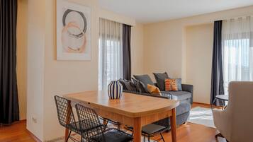 A modern living room with a wooden dining table, black wire chairs, a grey sofa with cushions, and a beige armchair.