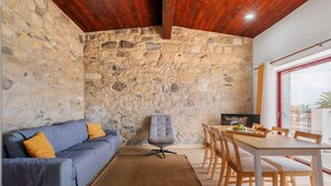 A room with a stone wall, a blue sofa with yellow cushions, a grey office chair, a wooden dining table with chairs, and a TV on the wall. - Casa Forno de Cal - By WeHostMadeira (Porto Santo)