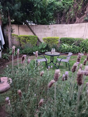 Un patio con una mesa redonda y dos sillas, rodeado de follaje y un muro bajo.