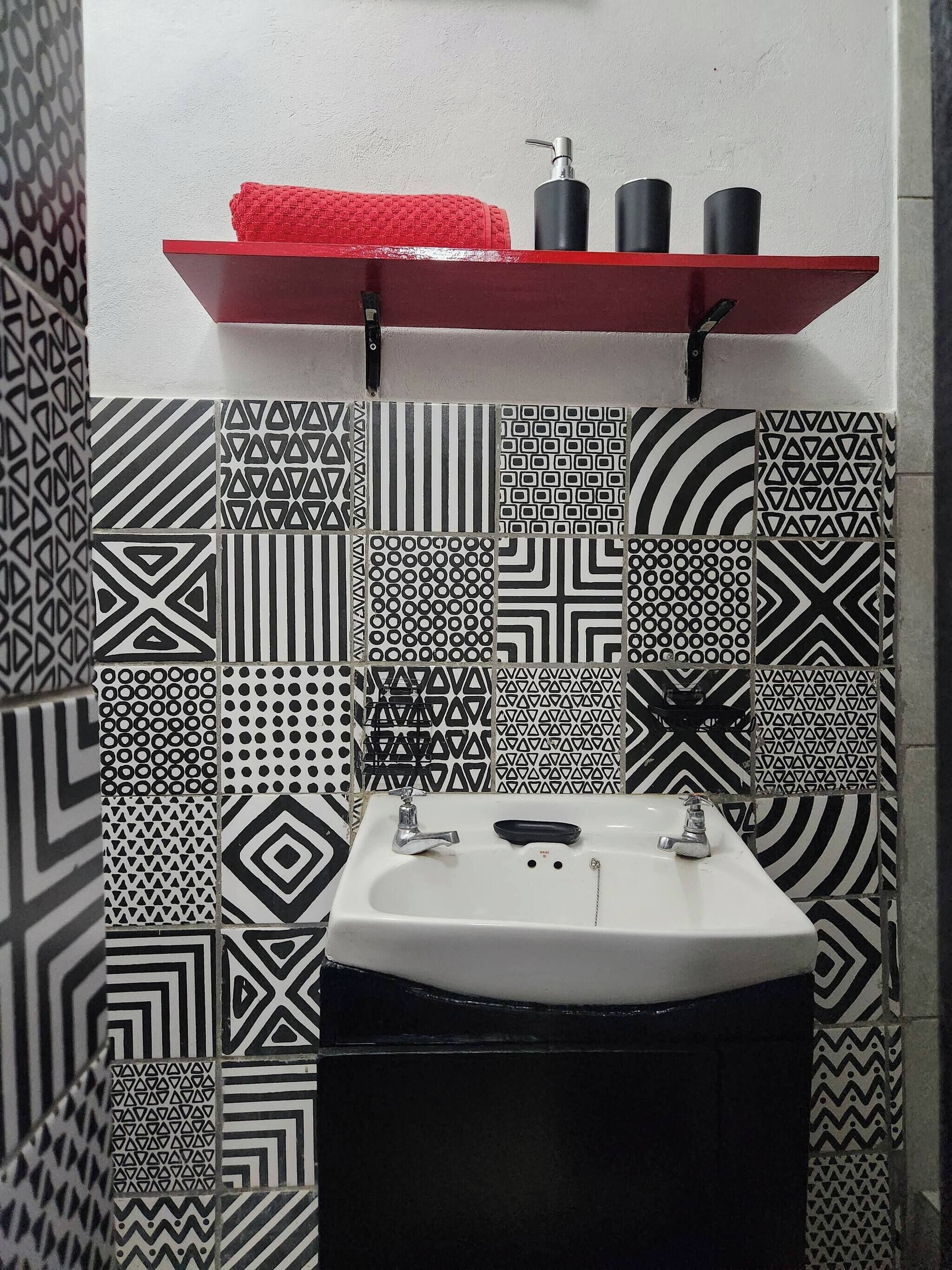Un baño con lavamanos blanco, azulejos con diseño blanco y negro, un sujetarropas rojo y artículos de aseo negro.