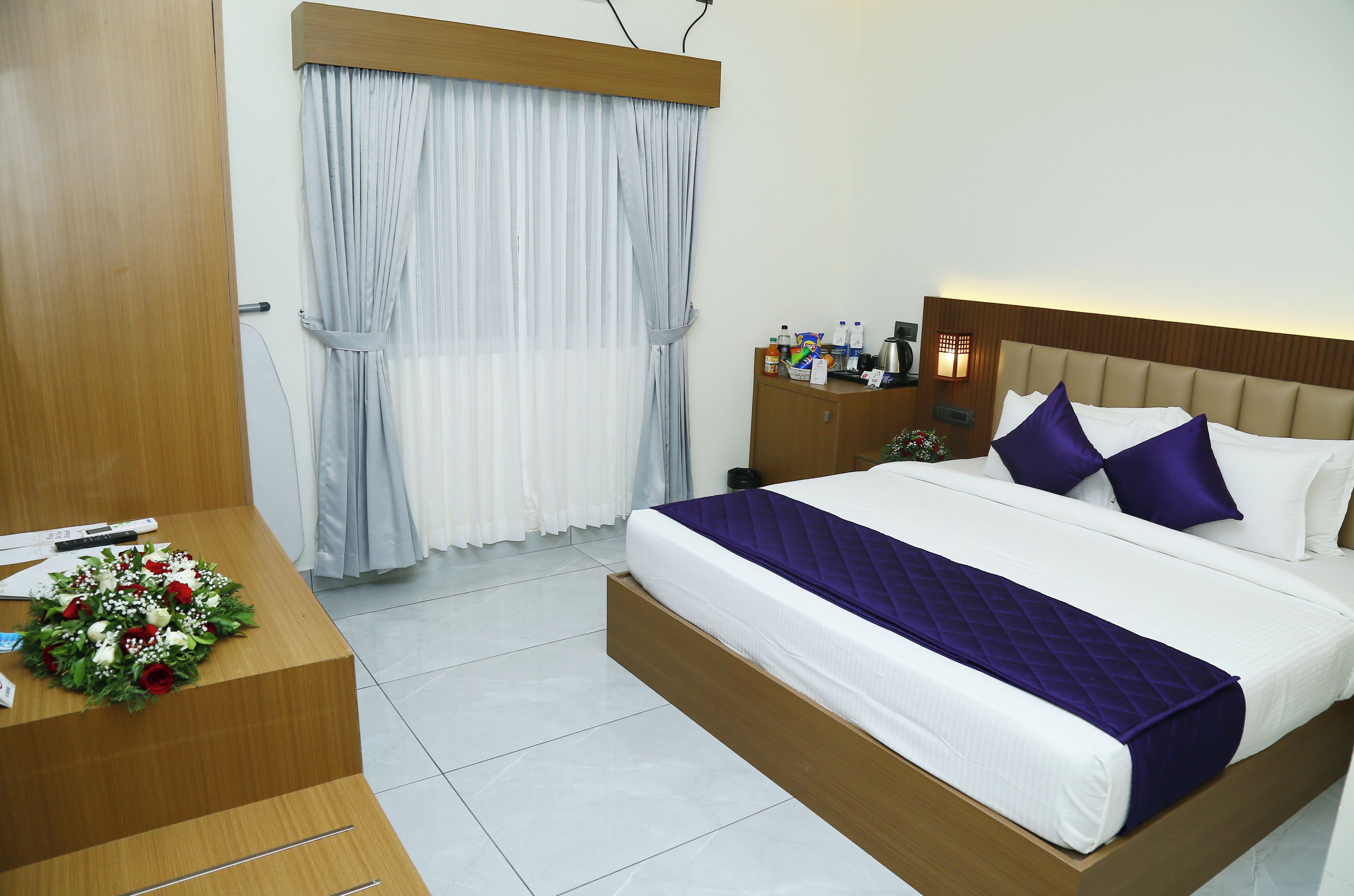 A hotel room with a bed, a desk, a nightstand, a window with curtains, and a wall-mounted lamp.