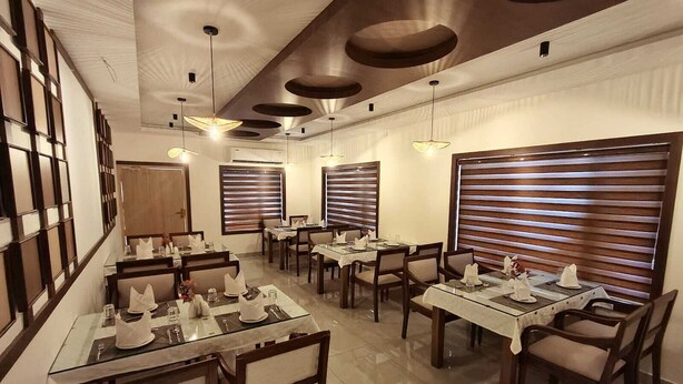 A dining area with multiple tables and chairs, ceiling lights, and large windows with blinds. - KABANI HERITAGE (Aluva)