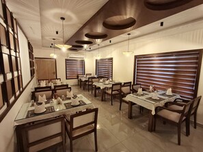 Un espace repas comprenant plusieurs tables et chaises, des luminaires au plafond et de grandes fenêtres équipées de stores.