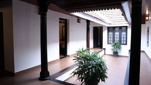 A corridor with a tiled floor, wooden columns, a potted plant, and a tiled ceiling with a decorative edge. - KABANI HERITAGE (Aluva)