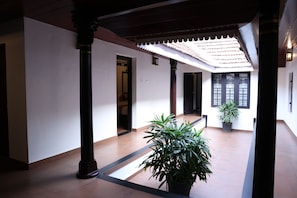 A corridor with a tiled floor, wooden columns, a potted plant, and a tiled ceiling with a decorative edge. - KABANI HERITAGE (Aluva)