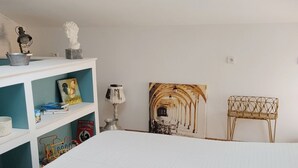 Une chambre à coucher avec un grand lit, une armoire blanche, une petite table avec une lampe, une chaise et une étagère murale où sont posés des livres et des objets décoratifs.