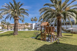 A playground with a slide and climbing structure, surrounded by palm trees and a building with balconies. - Appartement Don Pepe - Javea, Costa Blanca (Javea)