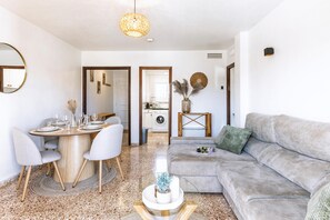 A modern living room with a grey sofa, a round dining table with chairs, a hanging light fixture, and a kitchen in the background. - Appartement Don Pepe - Javea, Costa Blanca (Javea)