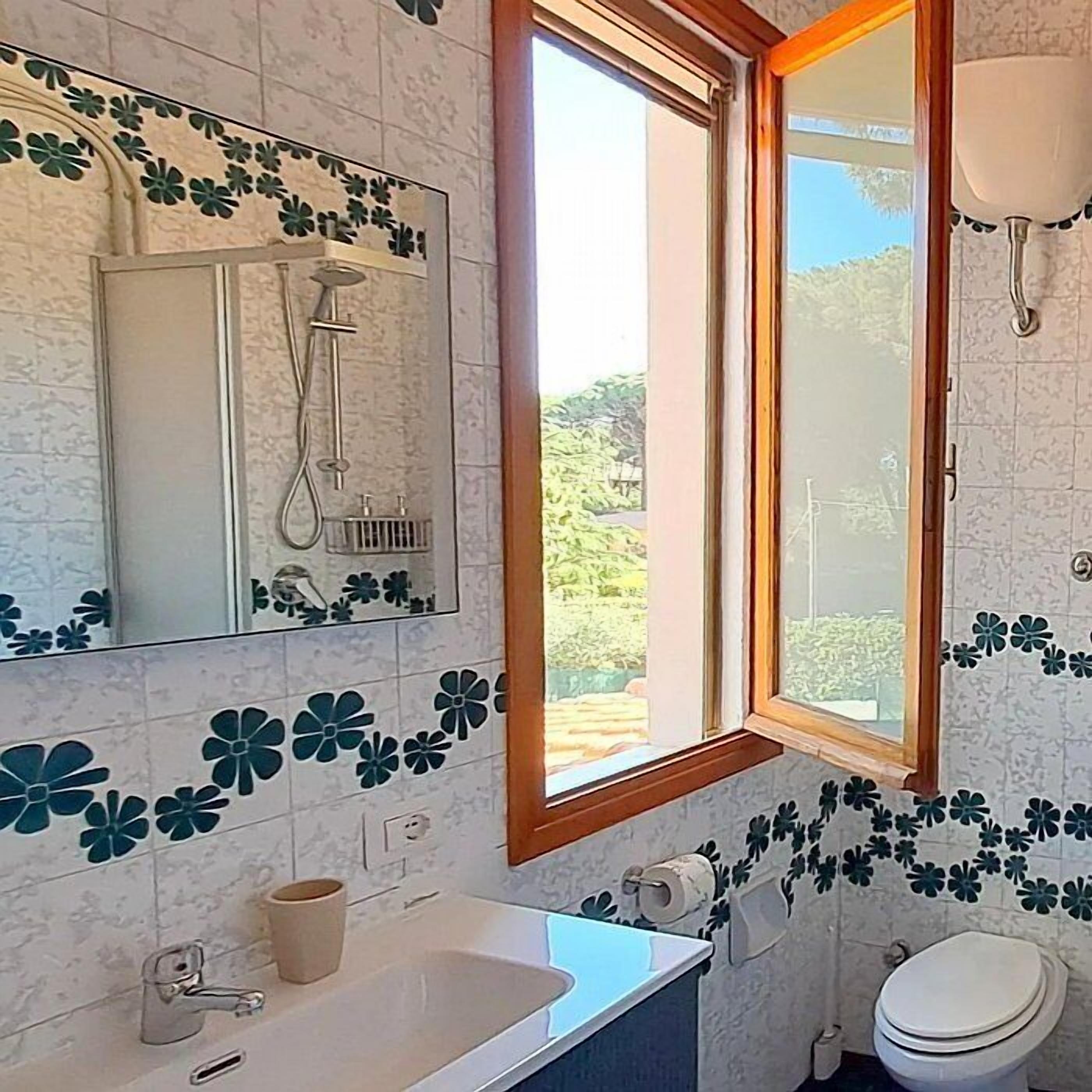 A bathroom with a shower, sink, toilet, and a window with a view of greenery.