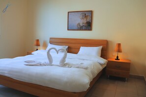 A neatly made bed with a heart-shaped towel arrangement, a wooden headboard, two bedside lamps, and a framed artwork on the wall.