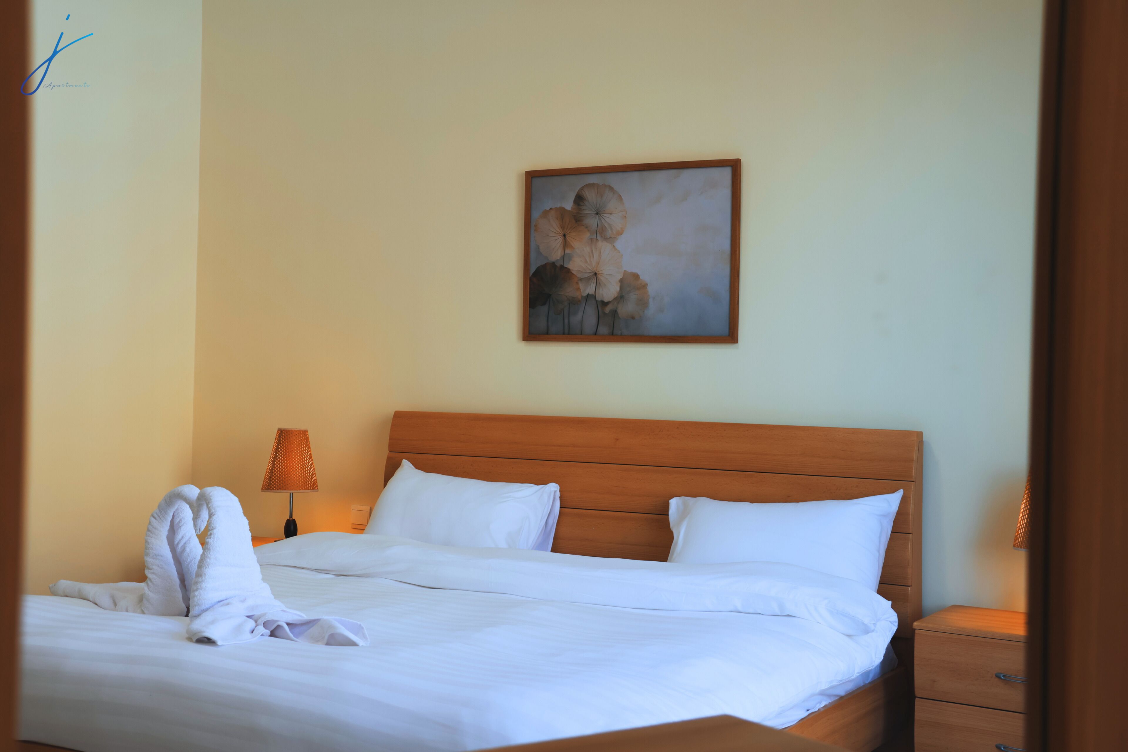 A neatly made bed with white linens, a wooden headboard, a nightstand with a lamp, and a painting on the wall.