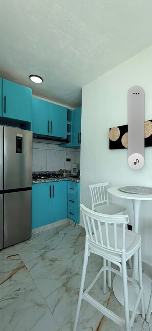 Una cocina con gabinetes color turquesa, un refrigerador de acero inoxidable, sillas blancas y una pequeña mesa redonda.