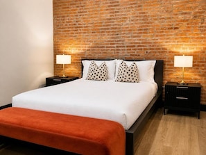 A bed with a white mattress and orange throw, two nightstands with lamps, and a brick wall. - Brick & Bed - Cheery Riverfront Loft Walk to Arch (St. Louis)