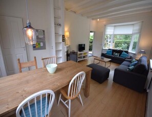 A dining area with a wooden table and chairs, a sofa, and a television in the background. - Seaside Escape With Large Garden (Oostkapelle)