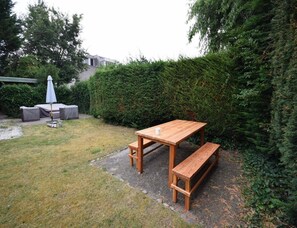 A wooden table with benches in a garden area with a hedge and a covered seating area. - Seaside Escape With Large Garden (Oostkapelle)