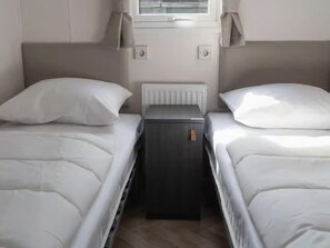 A bedroom with two beds, a small bedside table, a radiator, and a window with curtains. - Cozy Heinkenszand Retreat (Heinkenszand)
