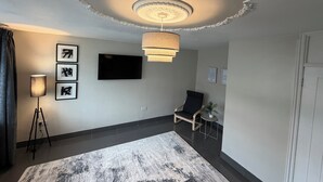 A modern living room with a flat-screen TV, a standing lamp, a chair, and a patterned rug. - 3 bedroom house, perfect for families, groups or contractors (Liverpool)
