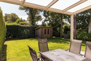 Eine überdachte Terrasse mit Tisch und Stühlen, ein Holzschuppen und ein gepflegter Rasen.