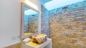 A bathroom with a stone wall, a white sink, a mirror, and a toilet. - Cocotoa Boutique Hotel and Villa (Gili Trawangan)