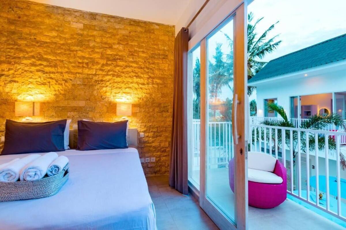 Ein Hotelzimmer mit Steinwand, einem Bett mit Kissen, einem Nachttisch und Blick auf einen Balkon mit Pool und Palmen.
